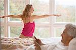 Woman practicing yoga warrior 2 pose and husband using digital tablet in bed