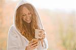Smiling woman in hooded bathrobe texting with cell phone and drinking coffee