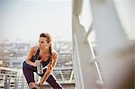 Female runner resting stretching leg and drinking water on sunny urban footbridge