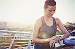 Female runner resting checking smart watch fitness tracker on sunny urban footbridge