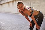 Tired fit female runner in sports bra with mp3 player armband resting on urban street