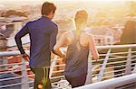 Runner couple running on sunny urban footbridge at sunrise