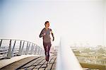 Female runner running on sunny urban footbridge at sunrise