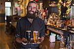 Portrait of young male bartender carrying tray of beer in public house