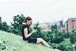 Young woman sitting on hillside reading smartphone texts above city