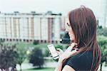 Young woman with long red hair holding smartphone and gazing above city