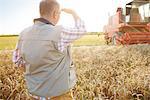 Rear view of farmer in wheat field looking at combine harvester