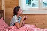 Young woman looking through window from log cabin bed, Tyrol, Austria