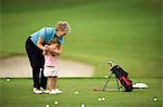 Senior woman teaching her young granddaughter how to swing a golf club on a golf course.