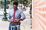 Young man standing beside bicycle, using smartphone, looking at wristwatch