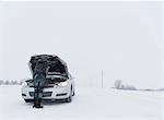 Rear view of man looking under hood of broken down car on snow covered landscape