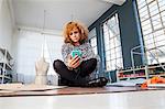 Mature female fashion designer cross-legged on workshop table looking at smartphone