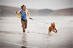 Smiling mid adult woman running along a beach with her dog.