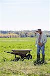 A young man in working clothes standing in a crop next to a wheelbarrow touching the brim of his cap.