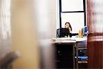 A young woman sitting at a desk in an office holding a telephone to her head.