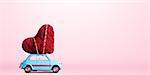 Blue retro toy car delivering craft heart for Valentine's day on pink background