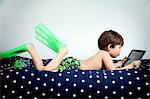 Boy lying on bed, wearing flippers and using digital tablet