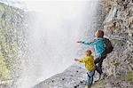 Mother and son, standing underneath waterfall, hands out to feel the water, rear view