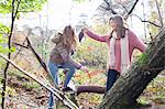 Teenage girl in forest helping frind to climb on fallen tree trunk smiling