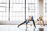 Full length front view of young women in gym legs open bending forward touching floor looking at camera