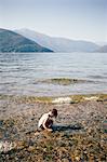 Side view of boy crouching in shallow water looking down and mountain range, Luino, Lombardy, Italy