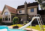 Rear view of a grey brick and cream cladding with grey trim cottage style home and landscaped backyard with in-ground swimming pool and slide in summer, Quebec, Canada