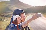 Young couple with backpacks hiking taking selfie with camera phone on sunny, remote road