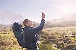 Young man with backpack hiking and using camera phone in sunny, remote field