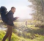 Young man with backpack hiking, checking smart phone in sunny field