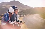 Young couple with backpacks hiking, resting at sunny, remote roadside with map and smart phone