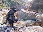 Young woman with backpack hiking, washing hands at sunny stream