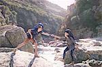 Young man helping girlfriend hiking, crossing sunny rocks in stream