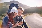 Young couple with backpack hiking, resting and using cell phone at sunny, remote roadside