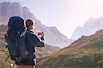 Young man with backpack using camera phone in sunny valley below mountains