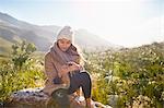 Young woman texting with cell phone on rock in sunny, remote field