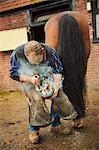 A farrier shoeing a horse, bending down and fitting a new horseshoe to a horse's hoof.  Steam from the hot metal.