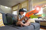 Young boy sitting cross legged on sofa, wearing virtual reality headset, holding laser guns, digital composite