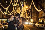 Couple taking smartphone selfie by Christmas lights at night, New York, USA