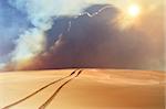 Vehicle tracks through desert and dunes leading into a sand, smoke and cloud filled sky. Digital photo manipulation.