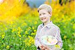 cheerful caucasian boy with basket full of easter eggs after egg hunt at beautiful field of blooming spring flowers