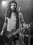 Photo of a young man with long hair and beard playing electric guitar on stage.