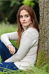 Outdoor portrait of beautiful thoughtful sad girl or young woman with red hair wearing a white jumper sitting & leaning against a tree in the countryside