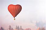Red air balloon in shape of heart fly above forest in foggy morning. Romantic postcard background on Valentine's Day. Adventure trip for couples. Sport and recreation travel theme. Nature background