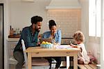 Girl Paints At Kitchen Table As Parents Look At Laptop