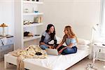 Two girls sitting on bed using laptop looking at each other