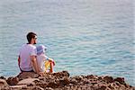 family of two, father and son, sitting on the rocks enjoying sunset at the tropical island