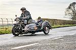 Senior man and grandson riding motorcycle and sidecar along rural road