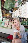 Mid adult woman with coffee cup making smartphone call at city sidewalk cafe