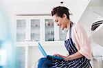 Young female baker sitting on kitchen counter typing on laptop