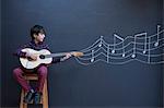 Boy playing guitar in front of chalkboard wall with showing musical notation